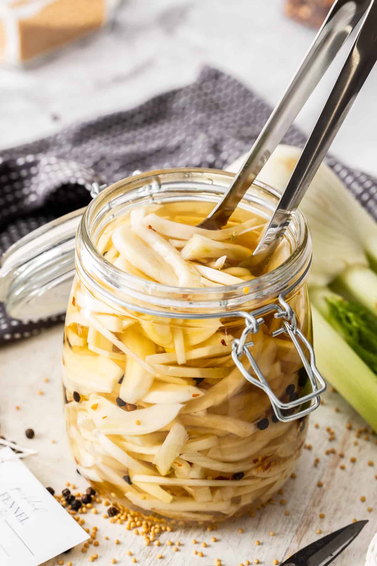 Jar of Pickled Fennel, sitting on a wooden board, with some tongs reaching in to pick up some pieces of fennel.