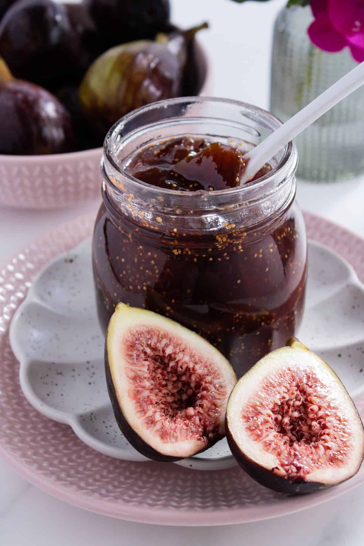 Open jar of Fig Jam, with a white spoon in it, sitting on a white saucer and pink plate with a fig cut in half on the edge.
