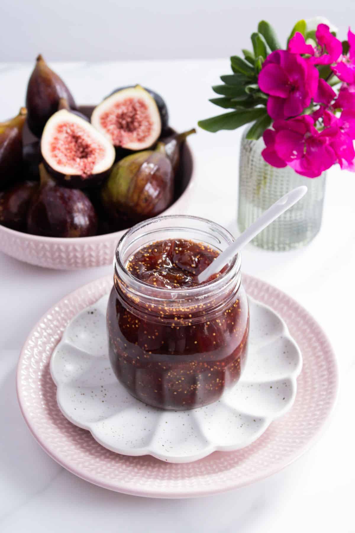 Open jar of Fig Jam, with a white spoon in it, sitting on a white saucer and pink plate, with a dish of figs and flowers behind.