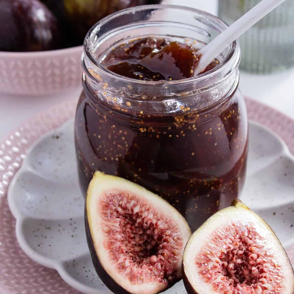 Open jar of Fig Jam, with a white spoon in it, sitting on a white saucer and pink plate with a fig cut in half on the edge.