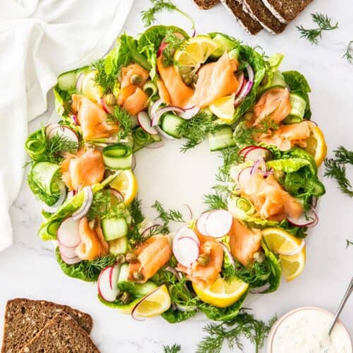 Smoked Salmon Wreath on a round white plate, sitting on a marble bench with some dressing and slices of rye bread around edge.
