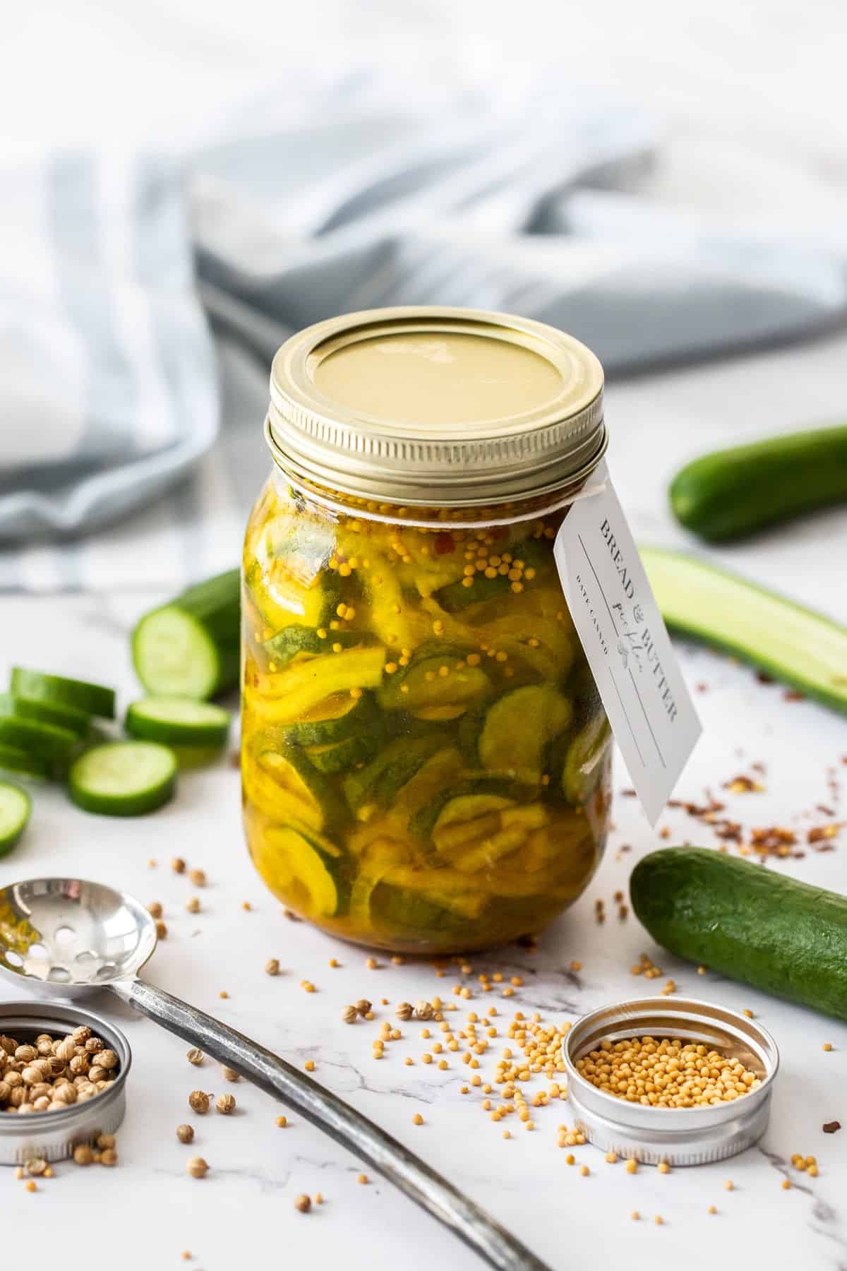 Jar of Bread and Butter Pickles, sealed with gold lid, and a label around the top, surrounded by spices and cucumber slices.