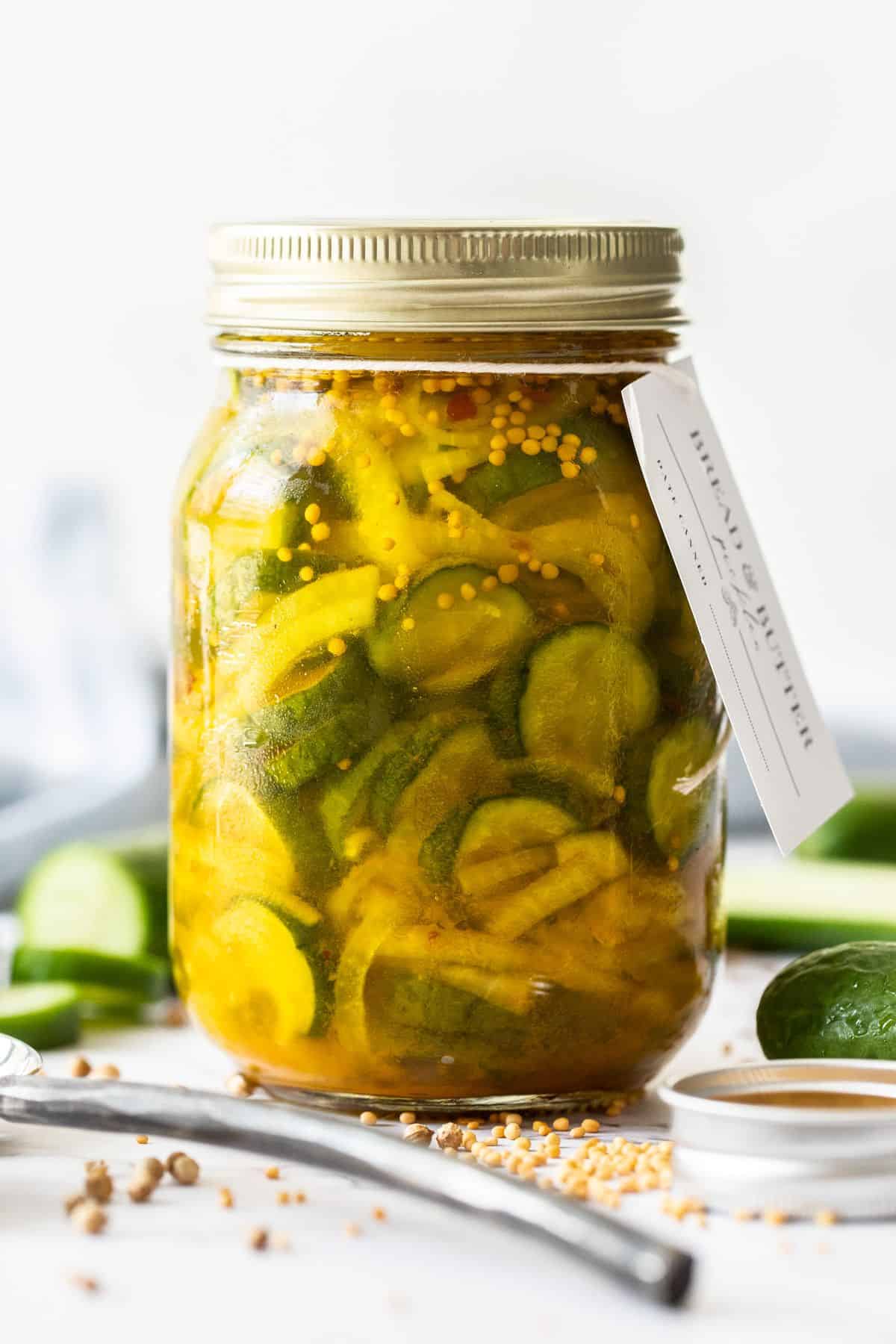 Jar of Bread and Butter Pickles, sealed with gold lid, and a label around the top, surrounded by spices and cucumber slices.