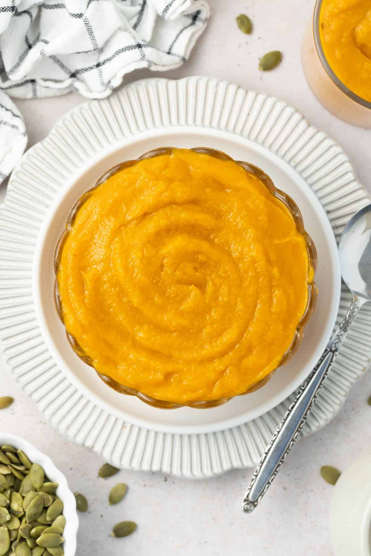 Round glass dish of pumpkin puree, sitting on a white plate, with a spoon on the edge.