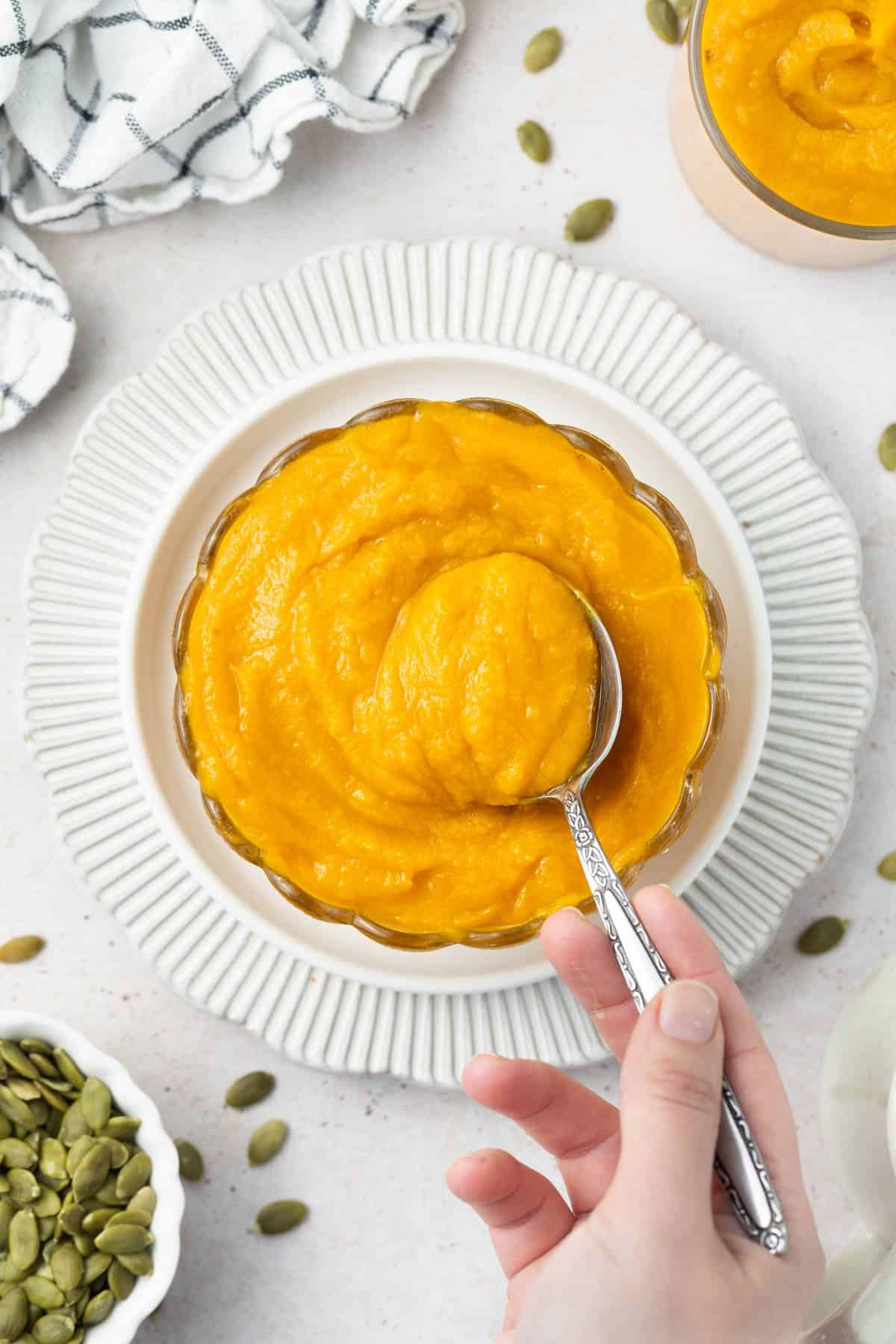 Round glass dish of pumpkin puree, sitting on a white plate, with a spoon dipping in to the puree.