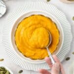 Round glass dish of pumpkin puree, sitting on a white plate, with a spoon dipping in to the puree.