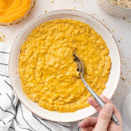 Round white bowl of pumpkin porridge with a spoon scooping up some of the mixture.