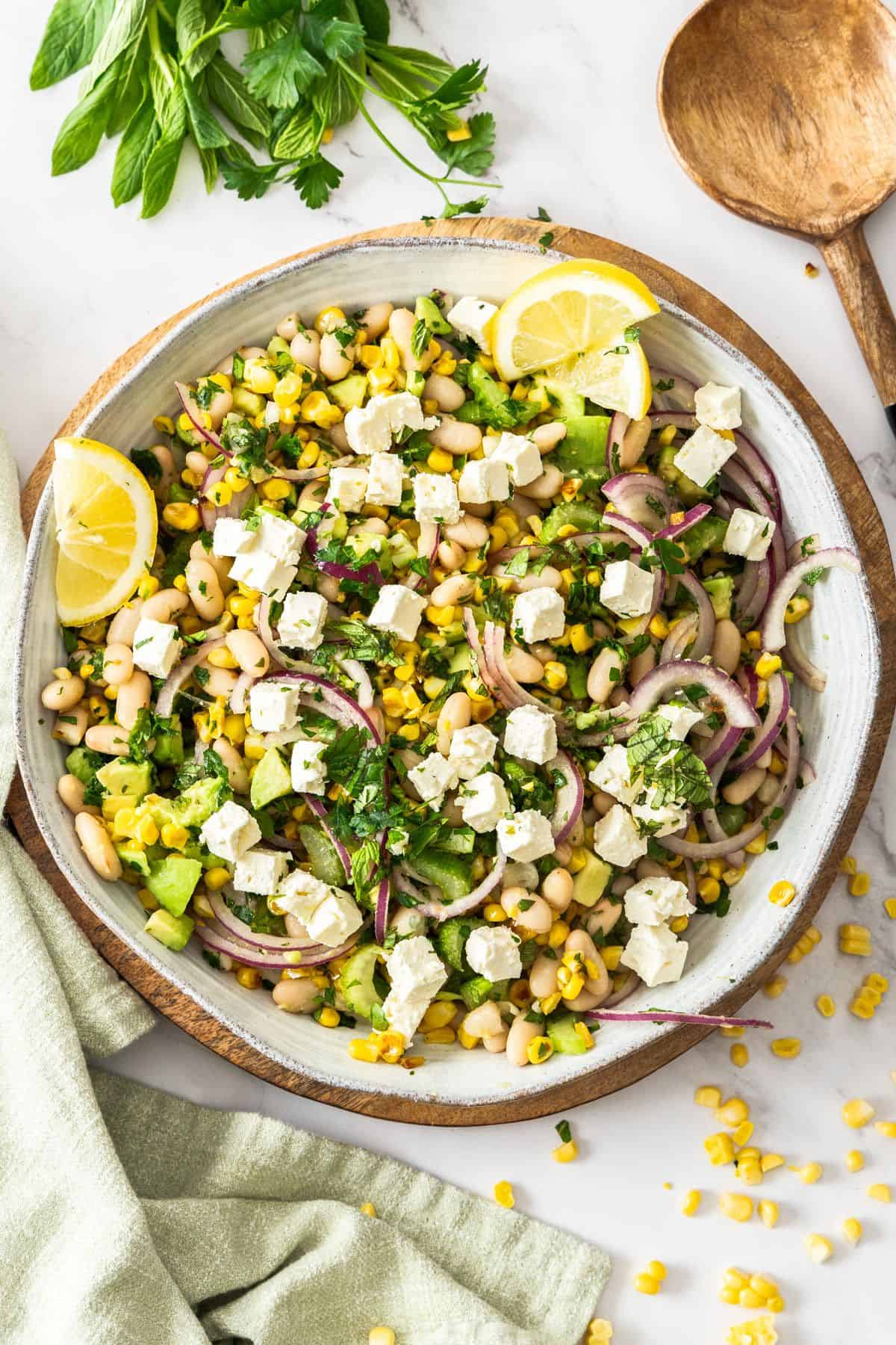 Round white bowl of Charred Corn Salad, with some lemon wedges on the side, and a wooden serving spoon.