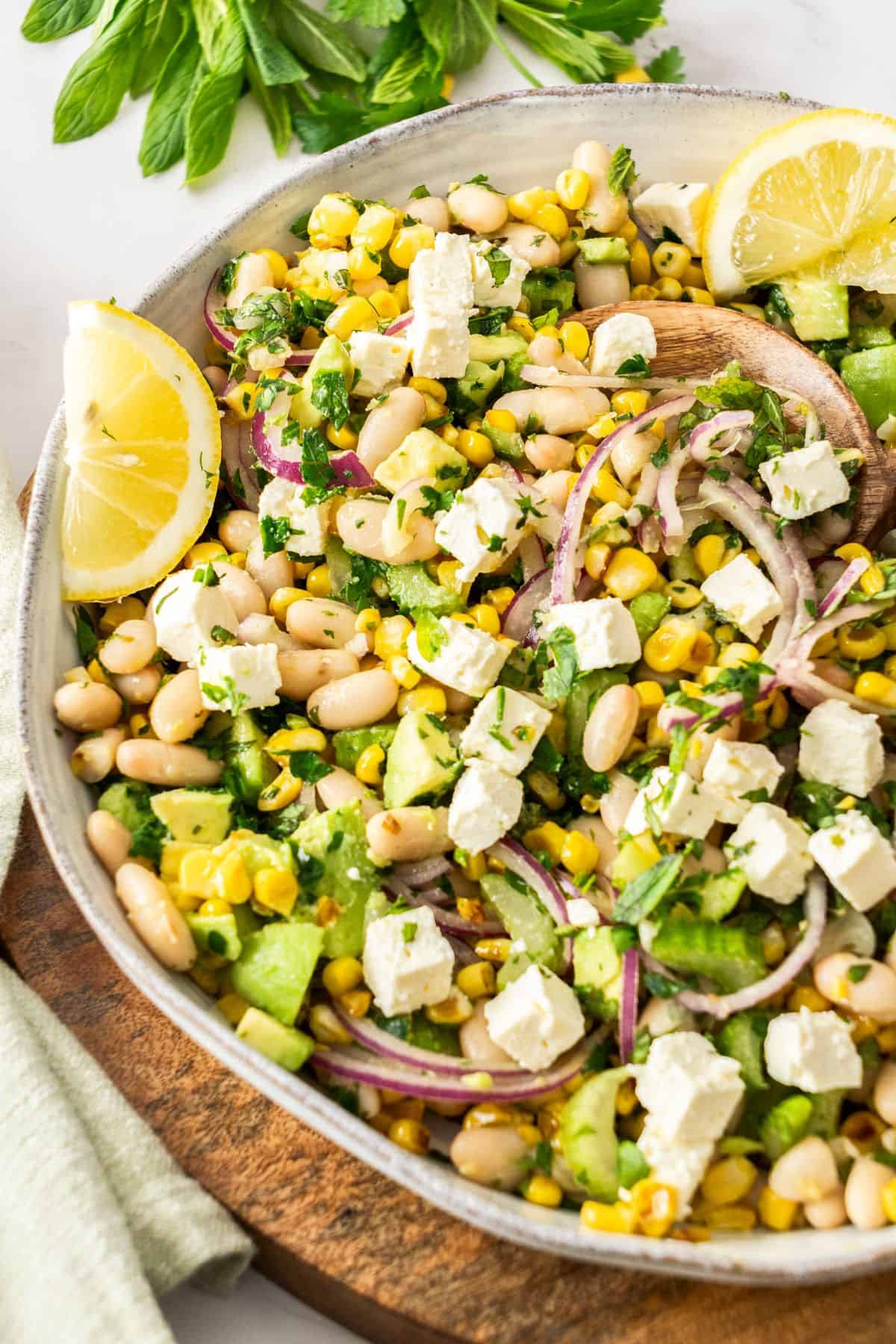 Round white bowl of Charred Corn Salad, with some lemon wedges on the side, sitting on a wooden board.