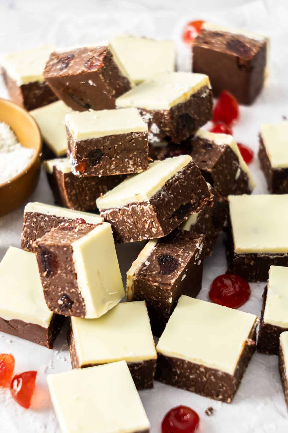 Cut squares of Dark Chocolate Cherry Fudge on some baking paper, with a dish of coconut and some glacé cherries scattered.