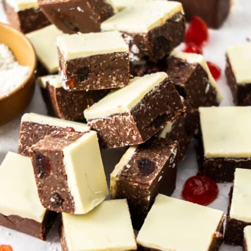 Cut squares of Dark Chocolate Cherry Fudge on some baking paper, with a dish of coconut and some glacé cherries scattered.