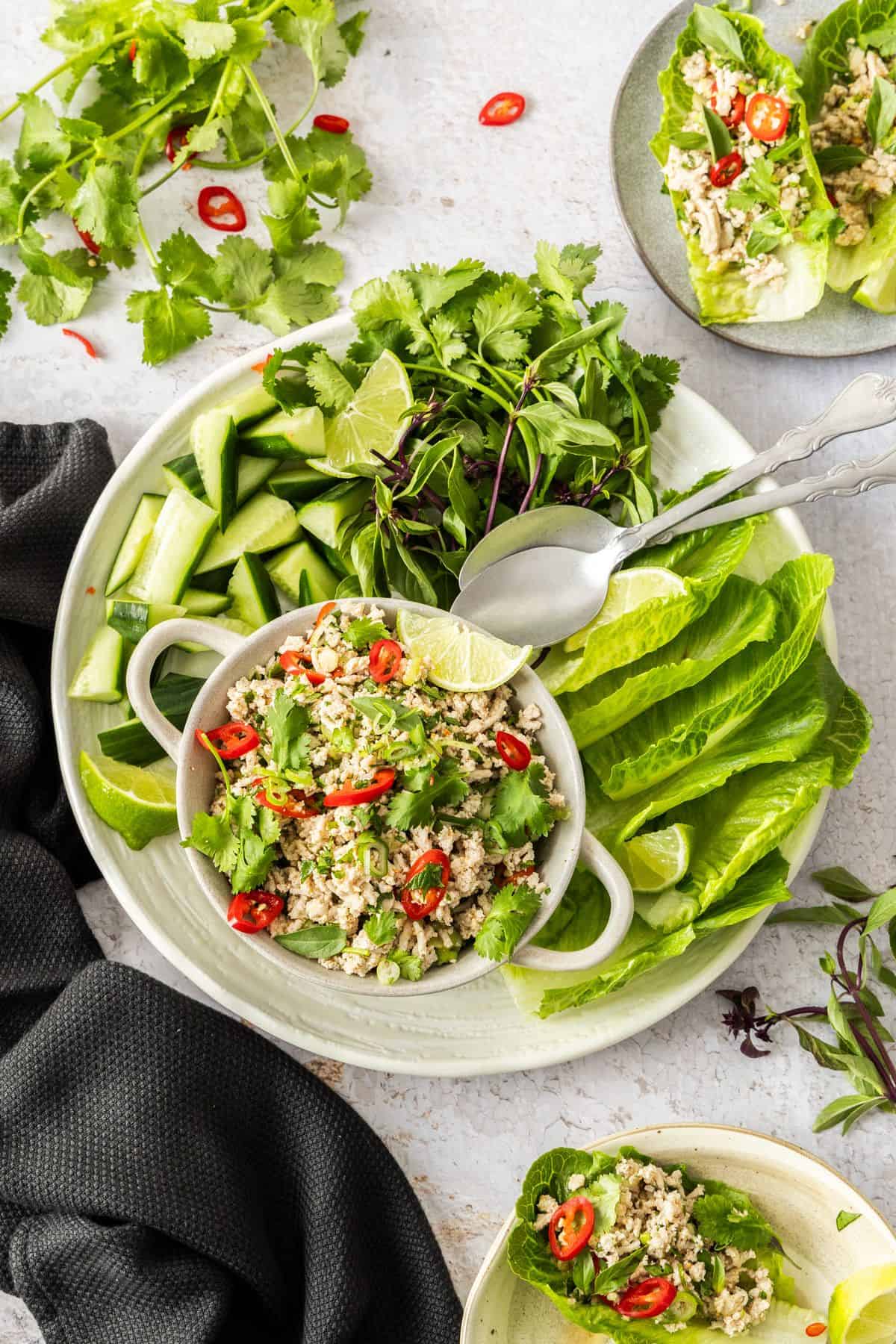 Bowl of Larb Gai, sitting on a round white plate with some lettuce, cucumber pieces, herbs and two silver spoons for serving.