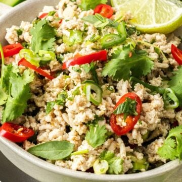 Up close shot of round white bowl of Larb Gai, garnished with pieces of chilli, herbs and lime wedges.