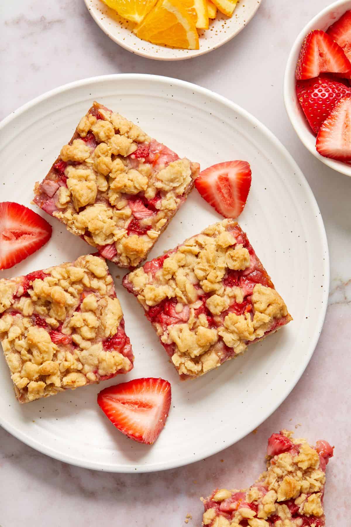 Three cut squares of Strawberry Oatmeal Bars on a round white plate, with some cut strawberry pieces.
