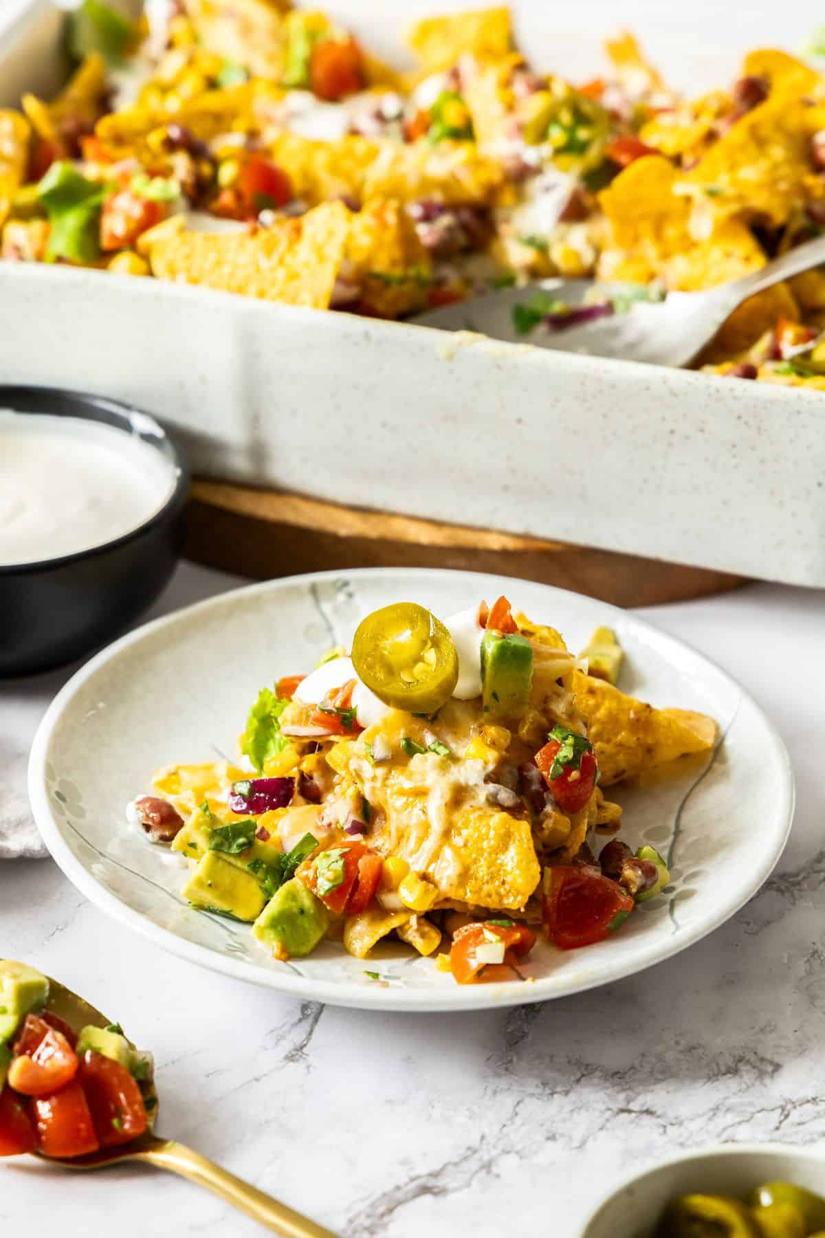 Small round white plate with a serving of Vegetarian Nachos, with the baking dish of nachos in the background.