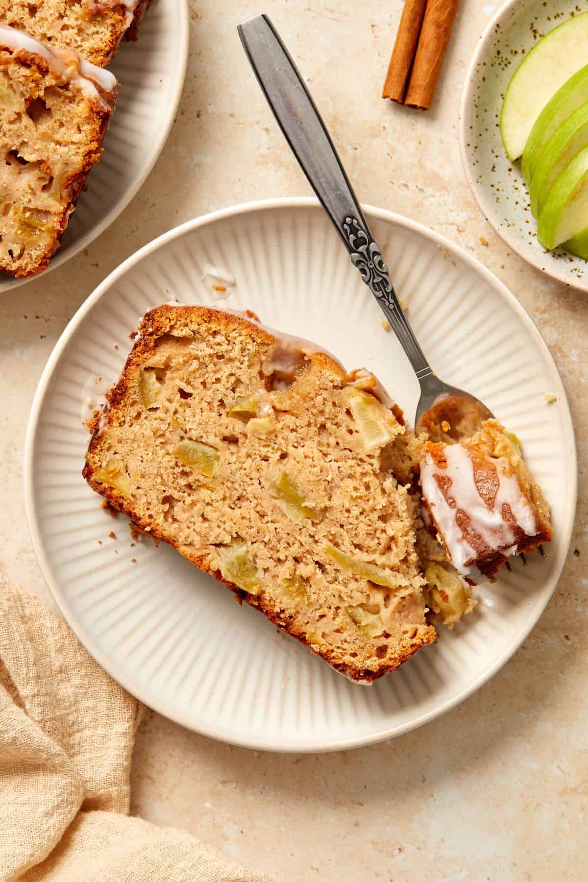A piece of Apple Loaf Cake on a small round white plate, with a fork cutting into the cake.