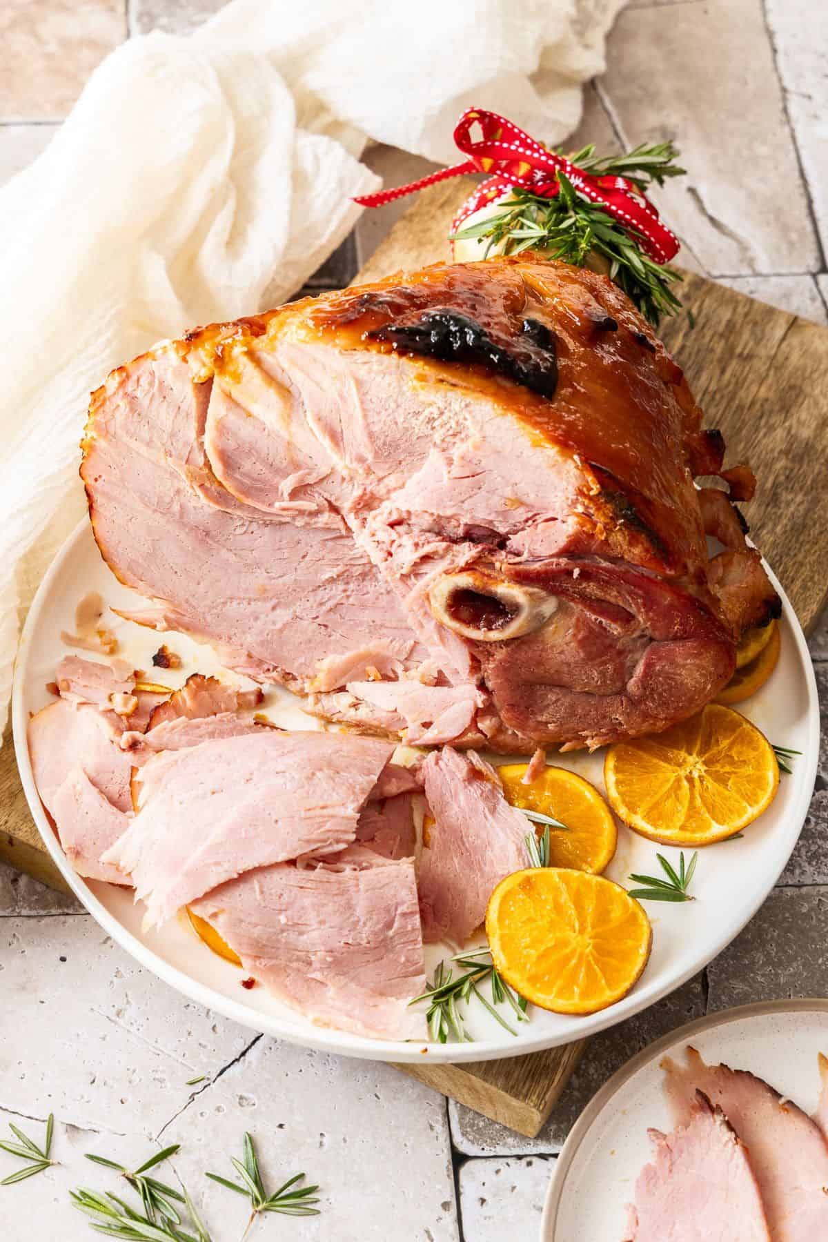 Marmalade Glazed Ham, sitting on a round white plate, with some slices of ham cut, and orange slices around the edge.