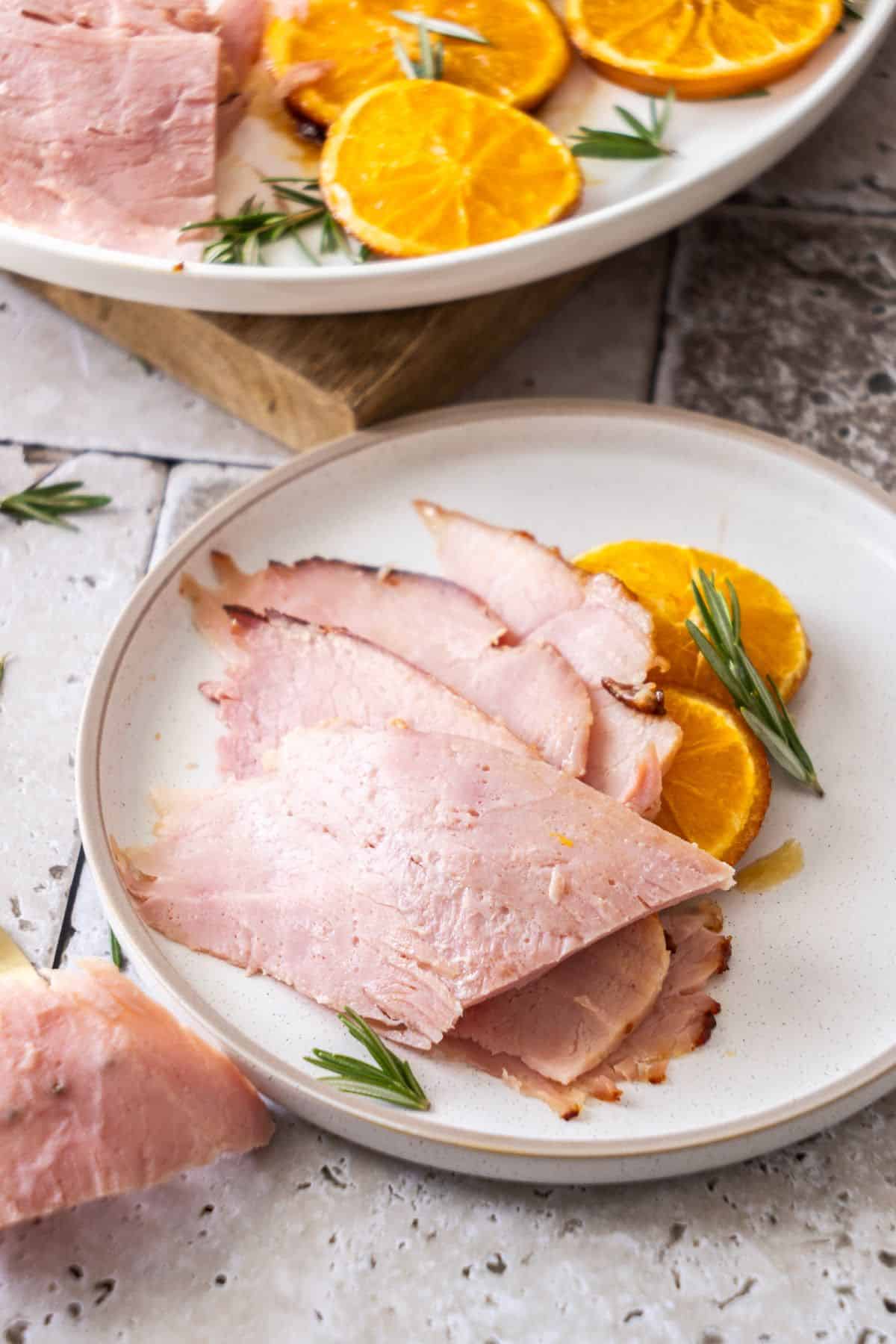 Slices of Marmalade Glazed Ham sitting on a small round white plate with some orange slices.
