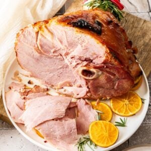 Marmalade Glazed Ham, sitting on a round white plate, with some slices of ham cut, and orange slices around the edge.