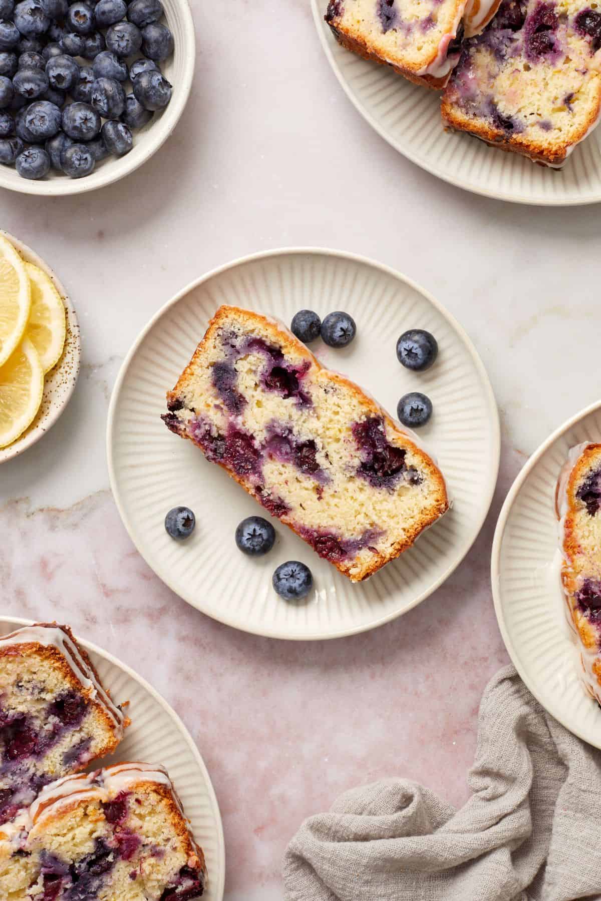 Slice of Blueberry Loaf Cake on a small round white plate, with some fresh blueberries around the edge.