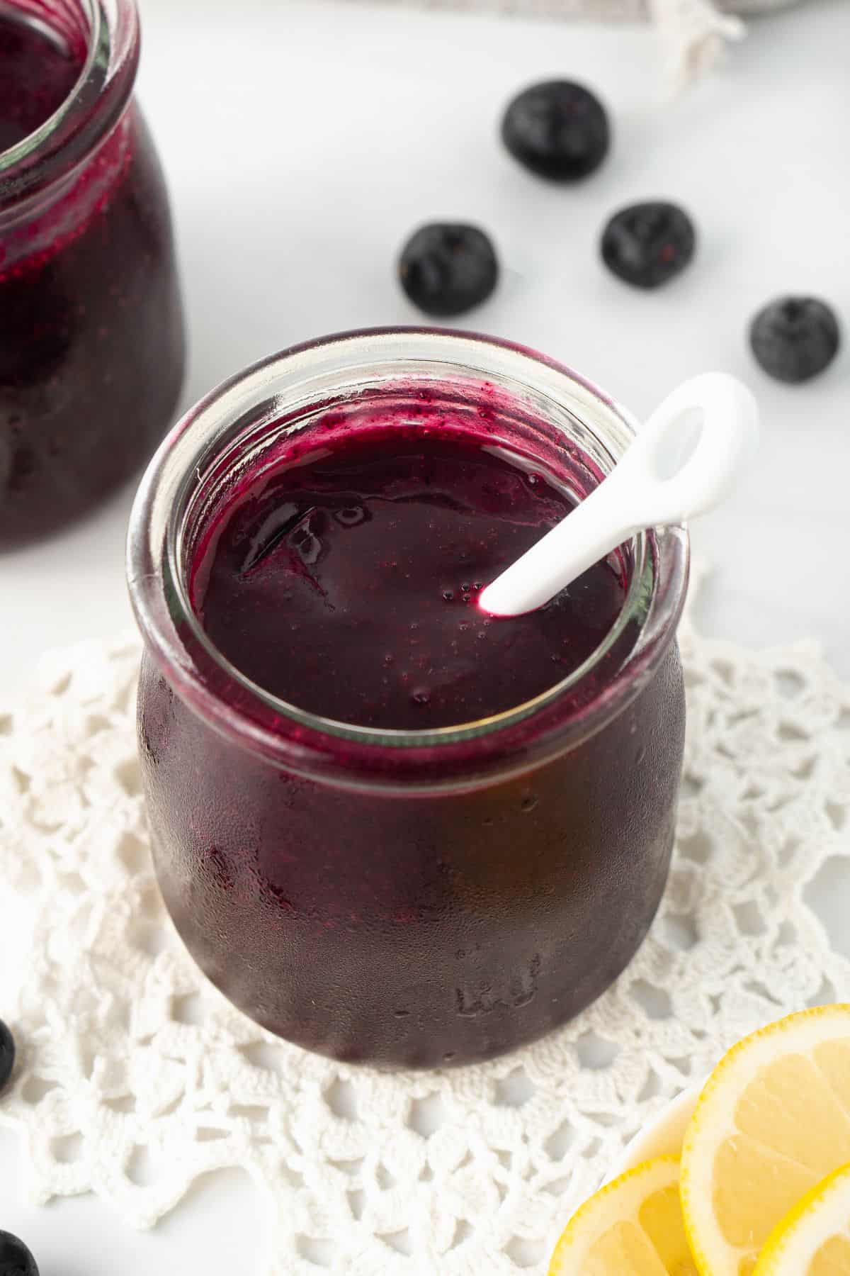 Two small jars of Blueberry Sauce, one with a spoon in it, surrounded by some blueberries and lemon slices.