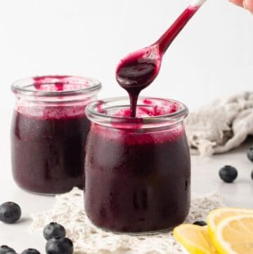 Two small jars of Blueberry Sauce, one with a spoon lifting sauce out of it, surrounded by some blueberries and lemon slices.