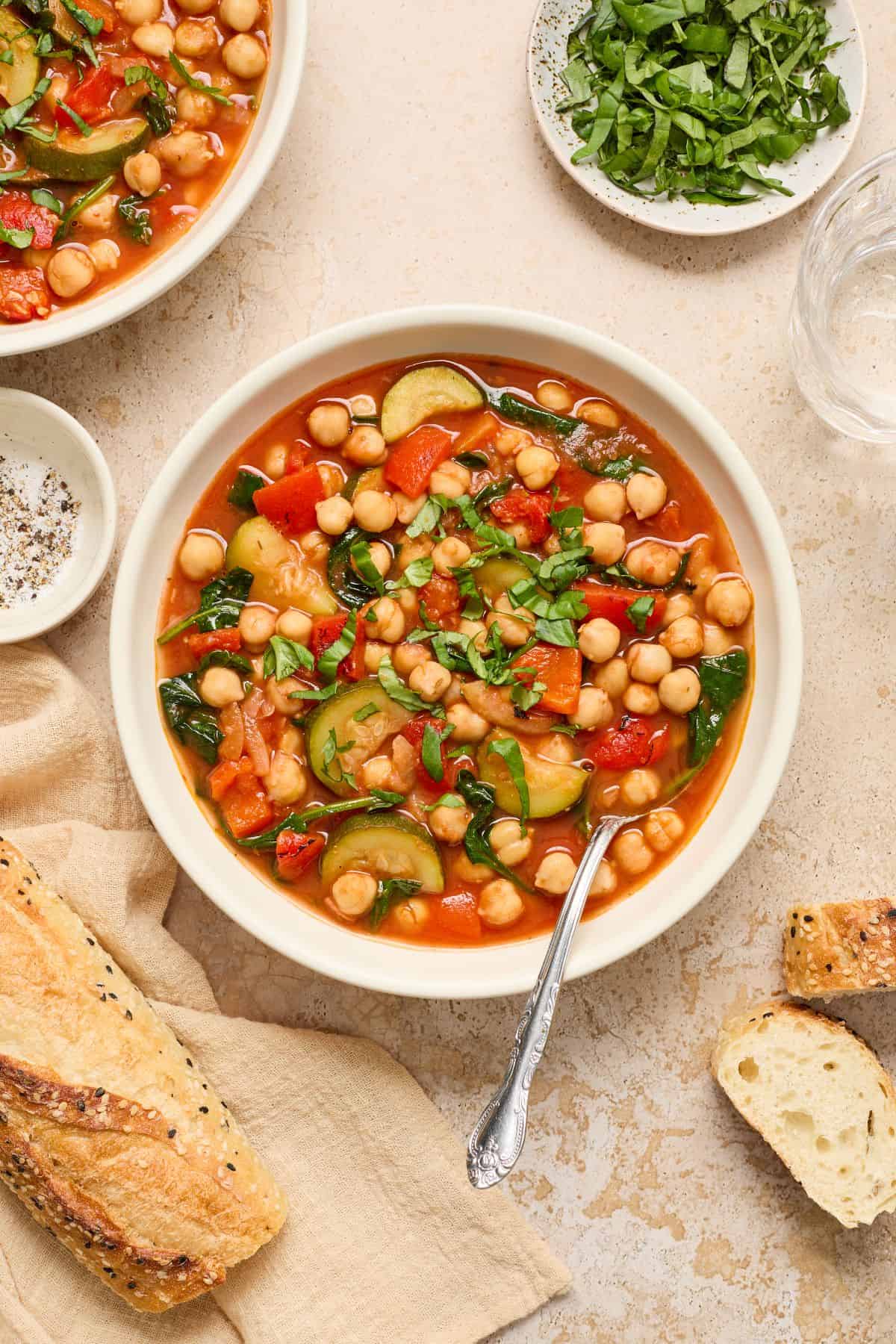 Two round white bowls of Chickpea and Zucchini Stew, one with a spoon resting in it, garnished with fresh basil.