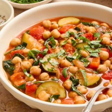 Round white bowls of Chickpea and Zucchini Stew with a spoon resting in it, garnished with fresh basil.