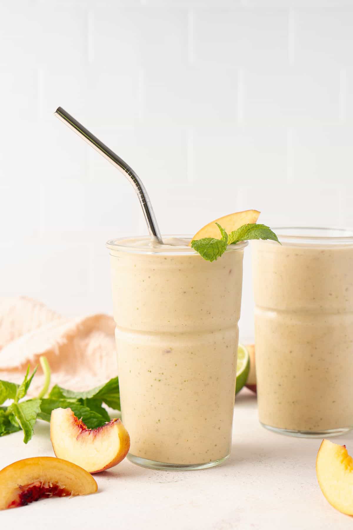 Two glasses full of Peach and Banana Smoothie, one with a straw, with some pieces of peach and fresh mint leaves on the edge.