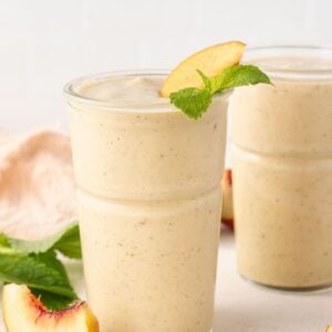 Two glasses full of Peach and Banana Smoothie, with some pieces of peach and fresh mint leaves on the edge.