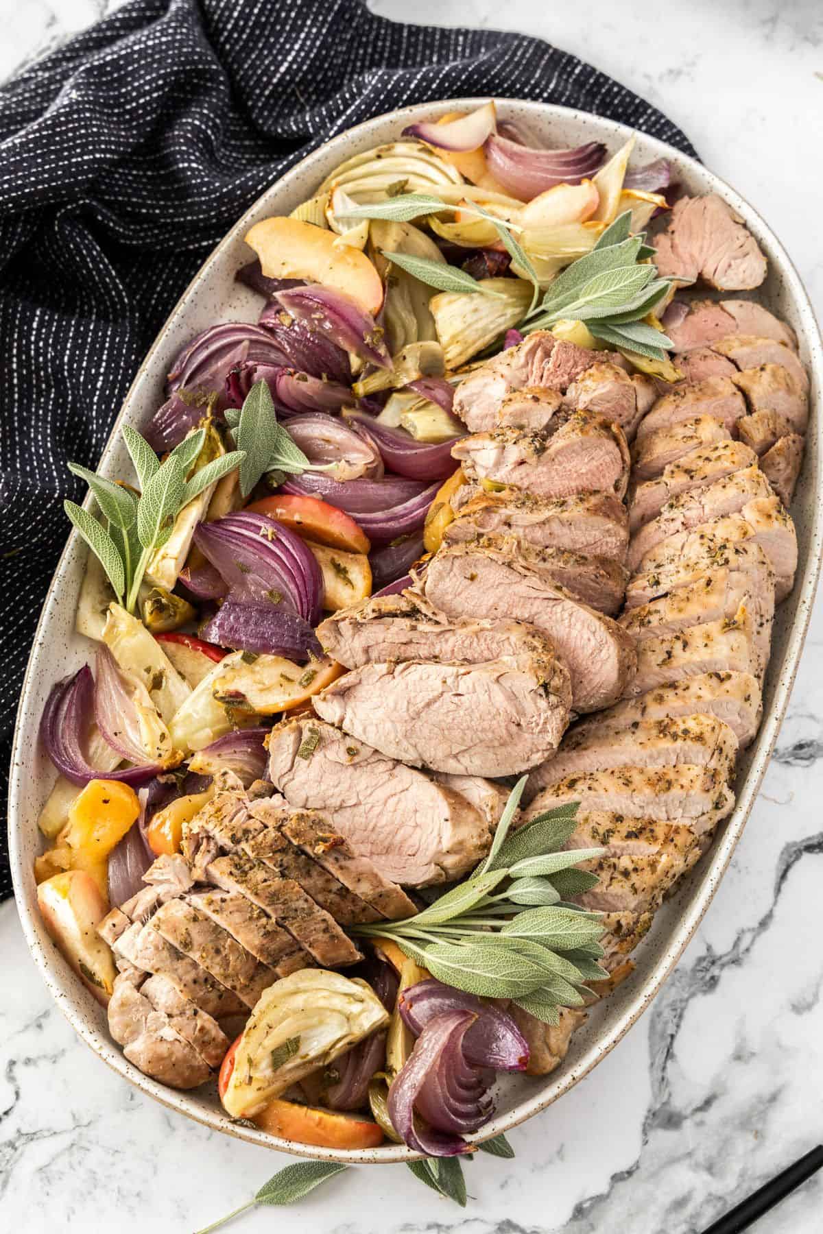 Platter of sliced Roasted Pork Tenderloin, surrounded by pieces of apple, fennel, onion and sage, with a black towel on edge.