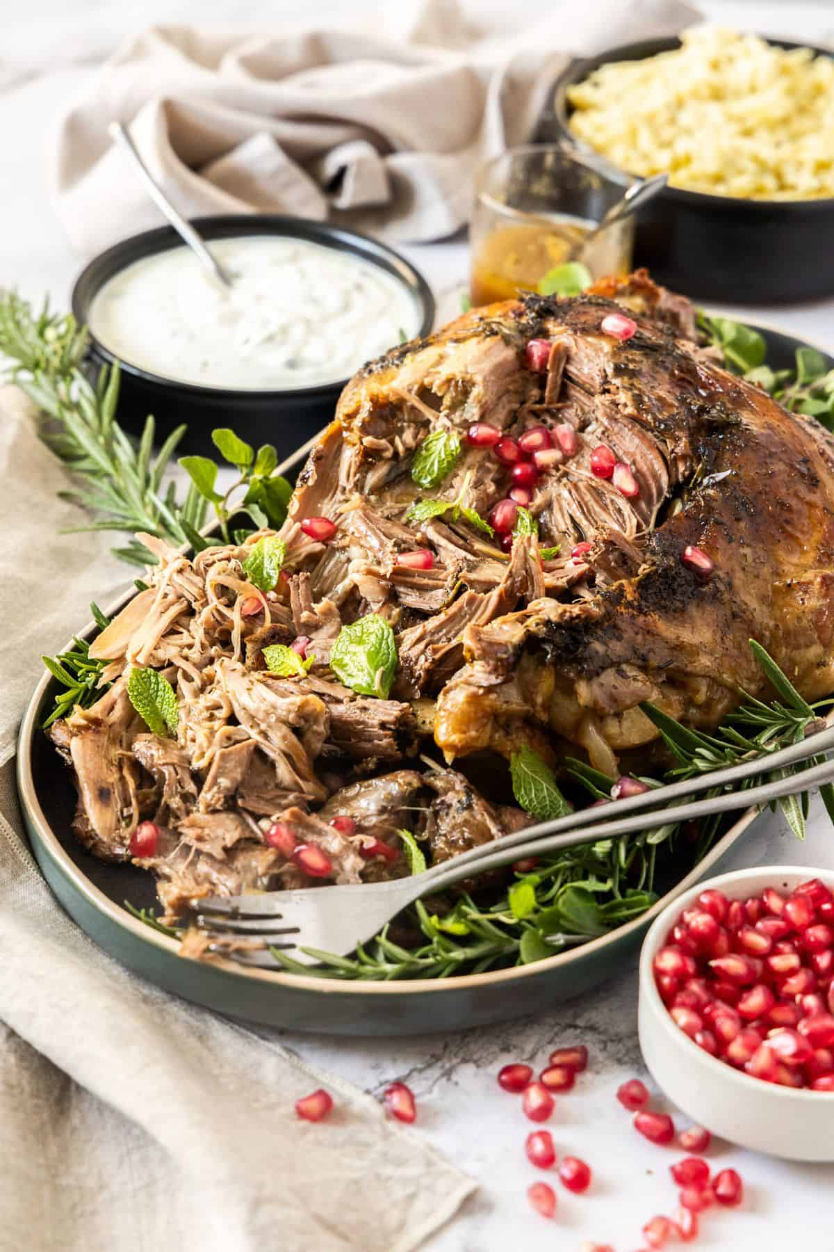 Slow Roasted Leg of Lamb, surrounded by fresh herbs and pomegranate, on a grey oval platter, with two forks on the edge.
