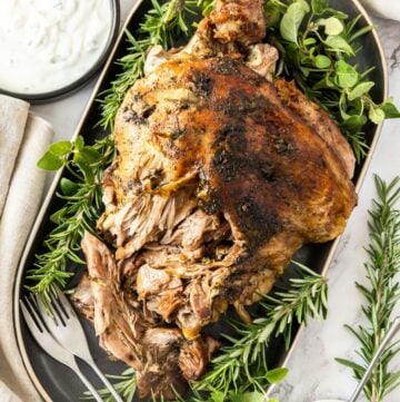 Slow Roasted Leg of Lamb, surrounded by fresh herbs, on a grey oval platter, with two forks on the edge.