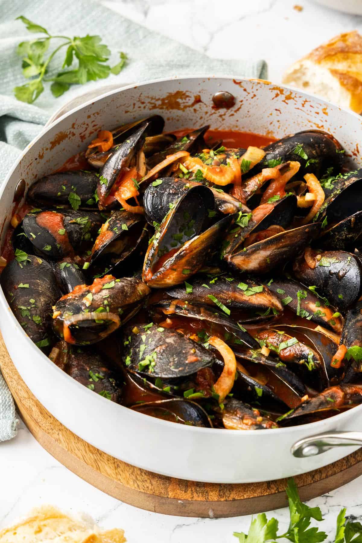 Frying pan with steamed mussels in tomato sauce, sprinkled with a parsley garnish, and some pieces of bread around the edge.