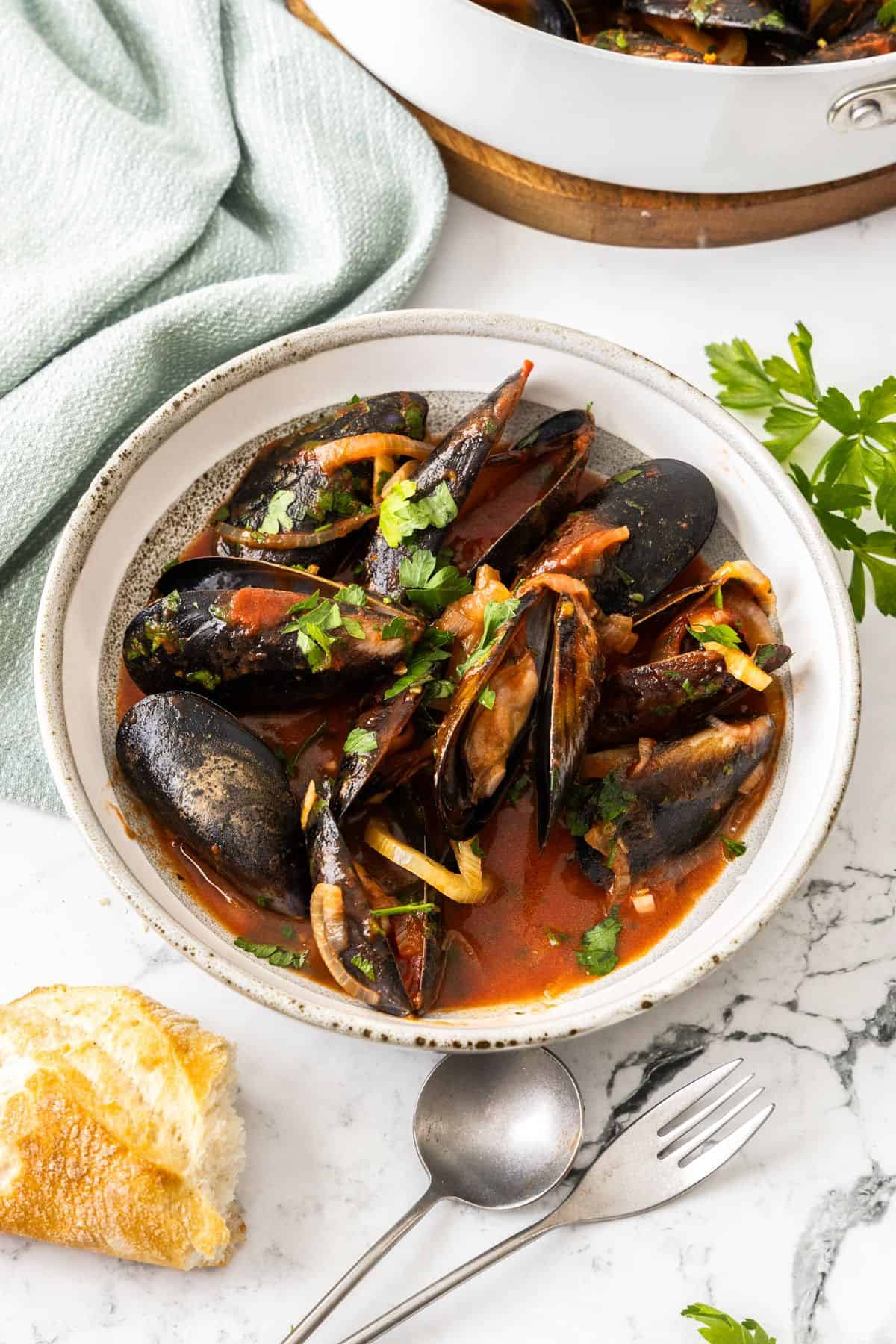 A serving of Mussels in Tomato Sauce, with some bread and cutlery sitting next to the bowl.