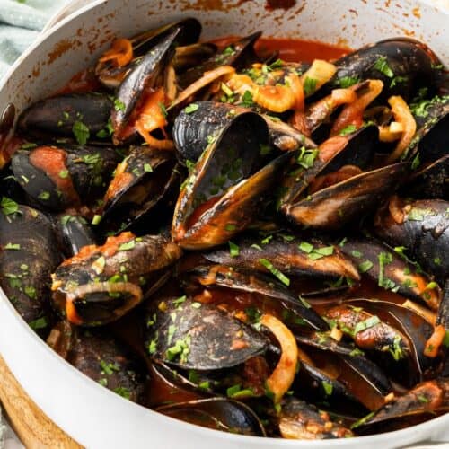 Frying pan with steamed mussels in tomato sauce, sprinkled with a parsley garnish, and some pieces of bread around the edge.