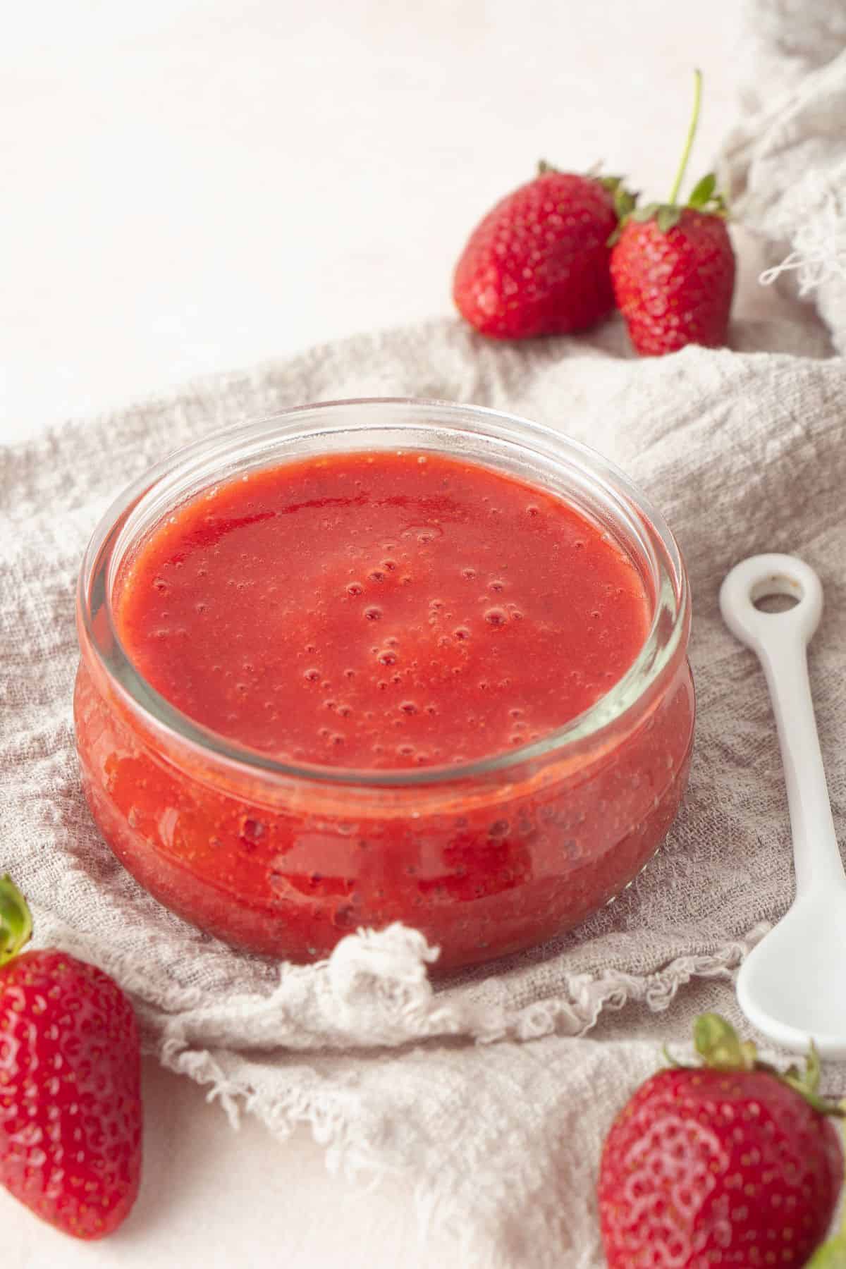 Small round glass dish of Strawberry Sauce, sitting on a cloth, with a white spoon and some strawberries around the edge.