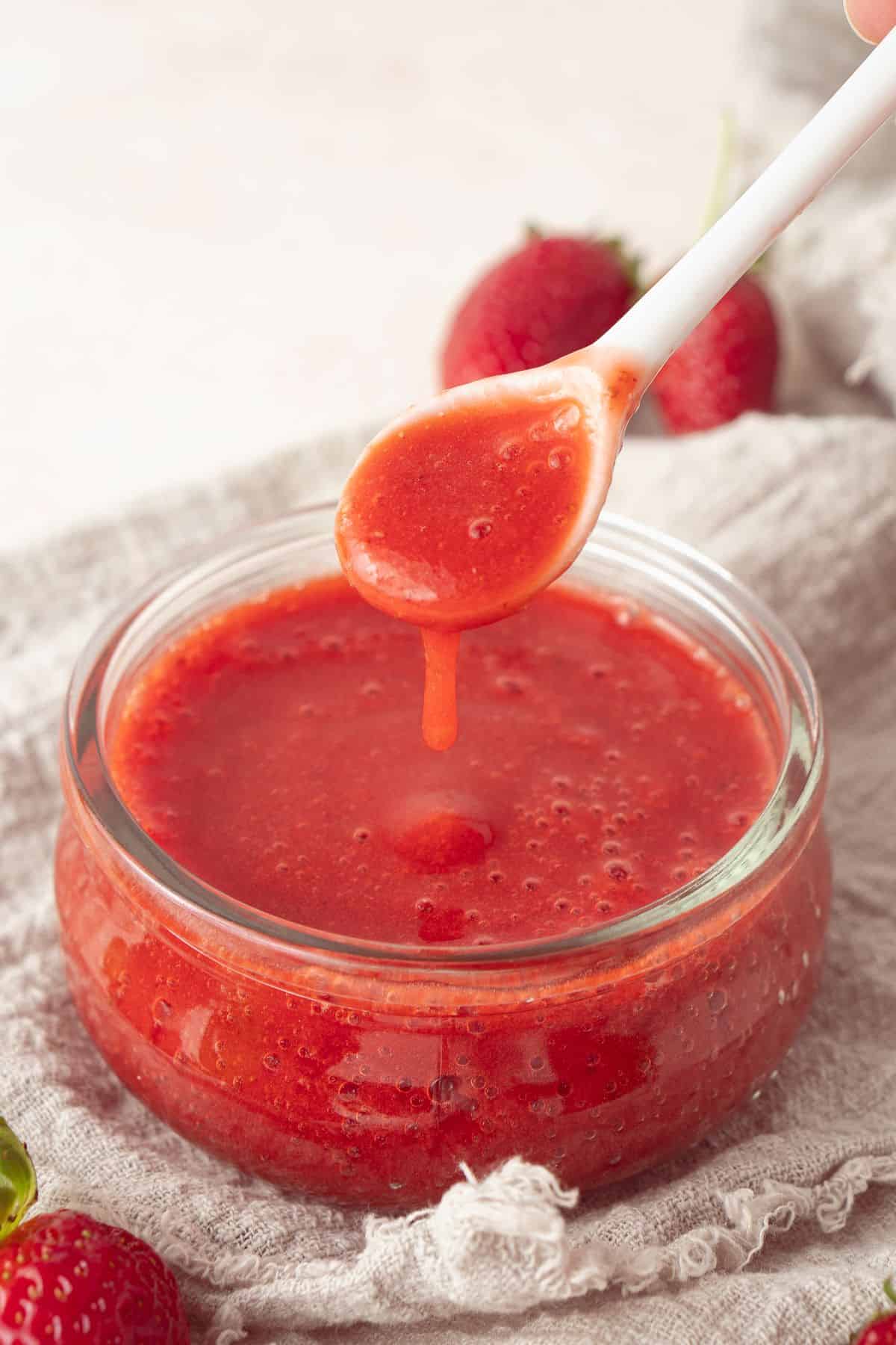 Small round glass dish of Strawberry Sauce, sitting on a cloth, with a white spoon lifting some sauce out of the dish.