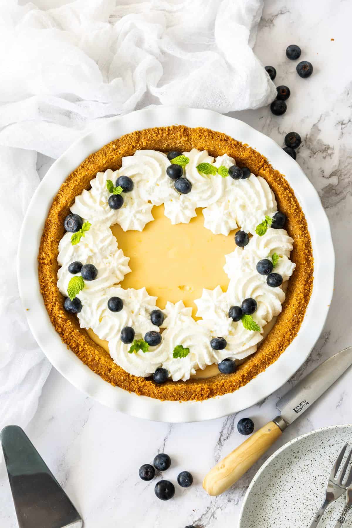 Creamy Lemon Pie in a round white pie dish, decorated with whipped cream, blueberries and mint leaves.