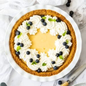 Creamy Lemon Pie in a round white pie dish, decorated with whipped cream, blueberries and mint leaves.