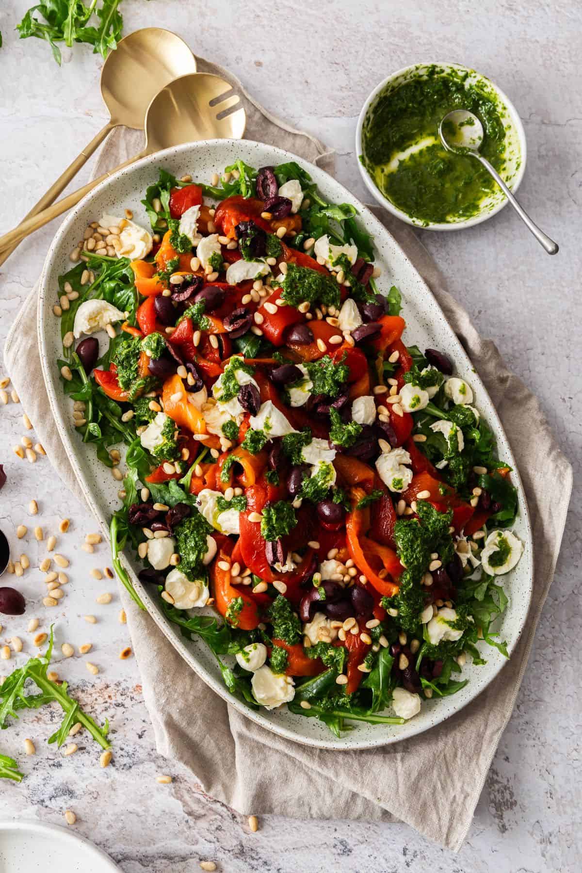 Oval platter with Roasted Capsicum (Red Pepper) Salad, with some serving utensils on the side.