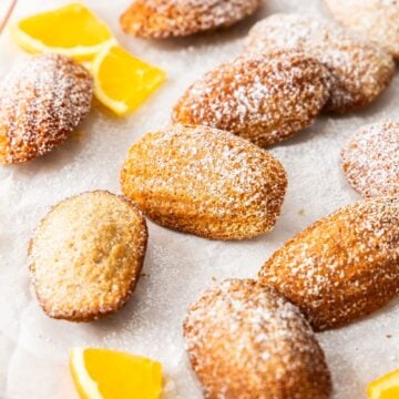 Spiced Orange Madeleines on some baking paper on a wooden board, dusted with icing sugar and a few pieces of fresh orange.