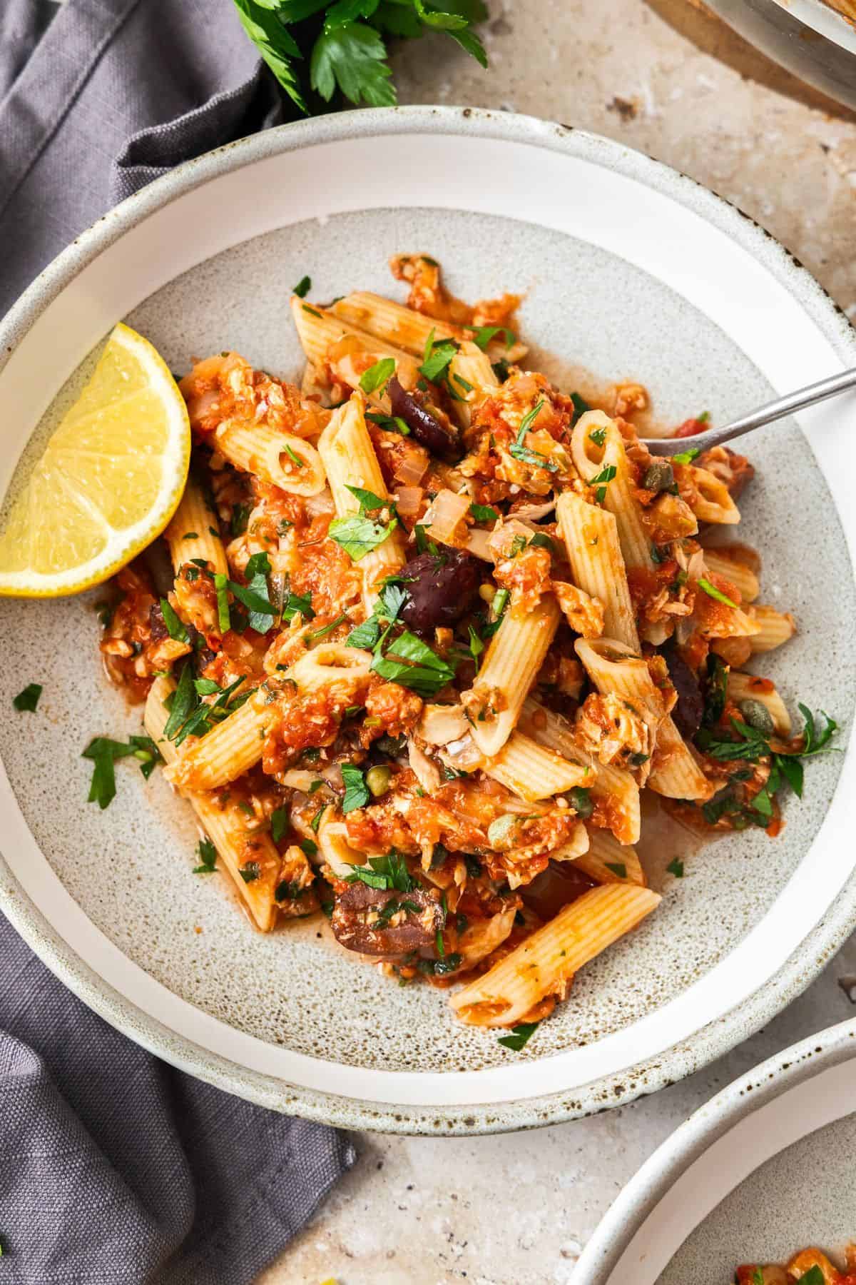 A serving of Spicy Tuna and Tomato Pasta in a round grey and white bowl, with a wedge of lemon on the edge.