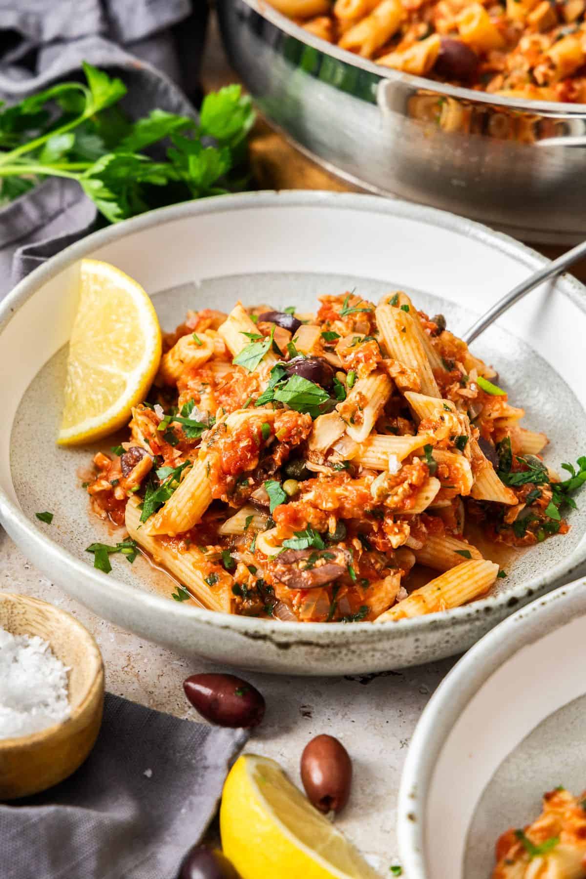 A serving of Spicy Tuna and Tomato Pasta in a round grey and white bowl, with a wedge of lemon on the edge.