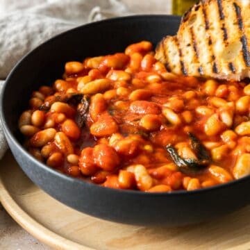 A serving of Tuscan Stewed Beans in a round black bowl, with a piece of grilled bread sitting on the beans.