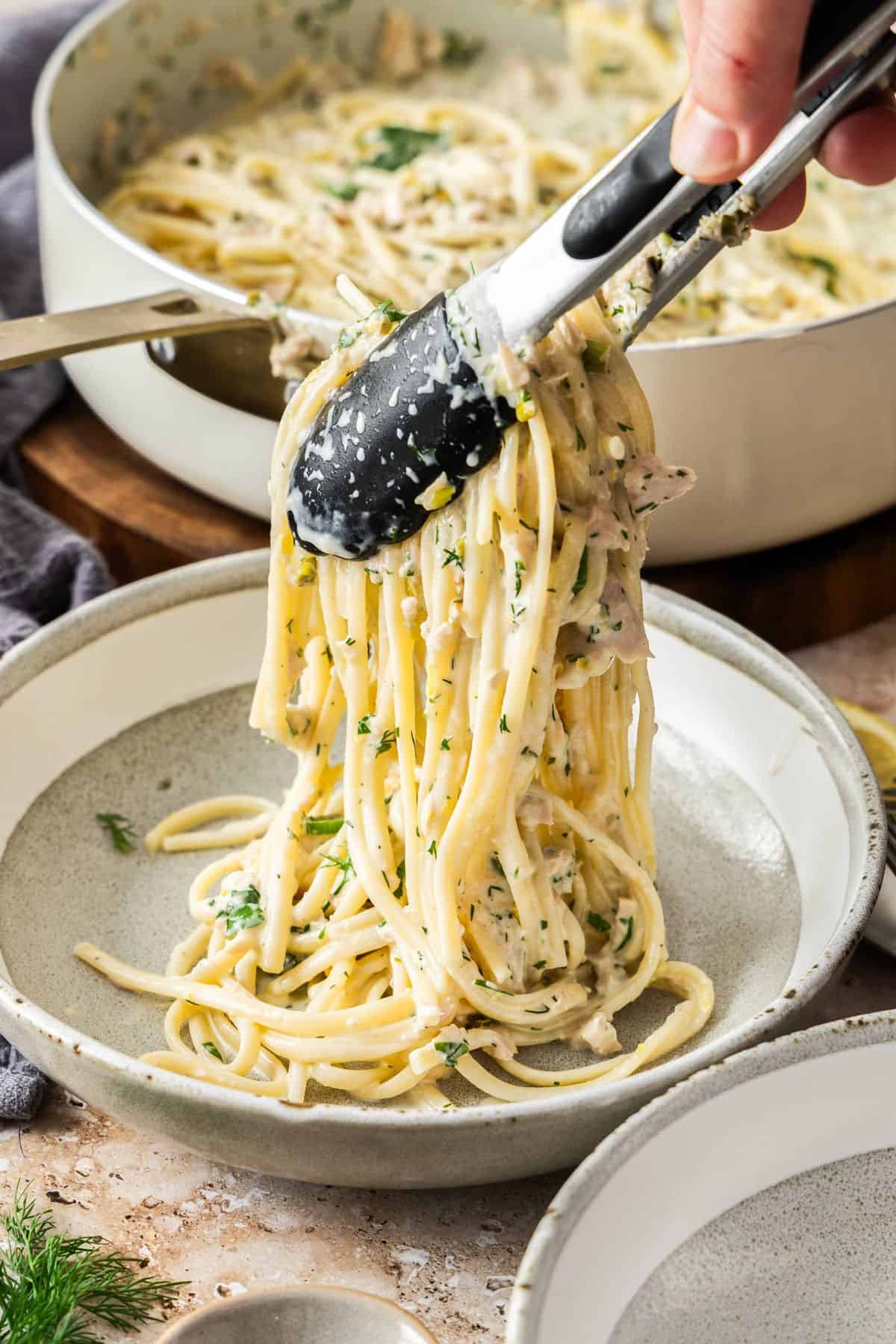 Tongs serving up some Creamy Tuna Pasta into a round grey and white bowl.