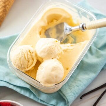 Plastic container of ice cream, with some serves scooped out, with some ice cream cones and a bowl of strawberries on the edge.