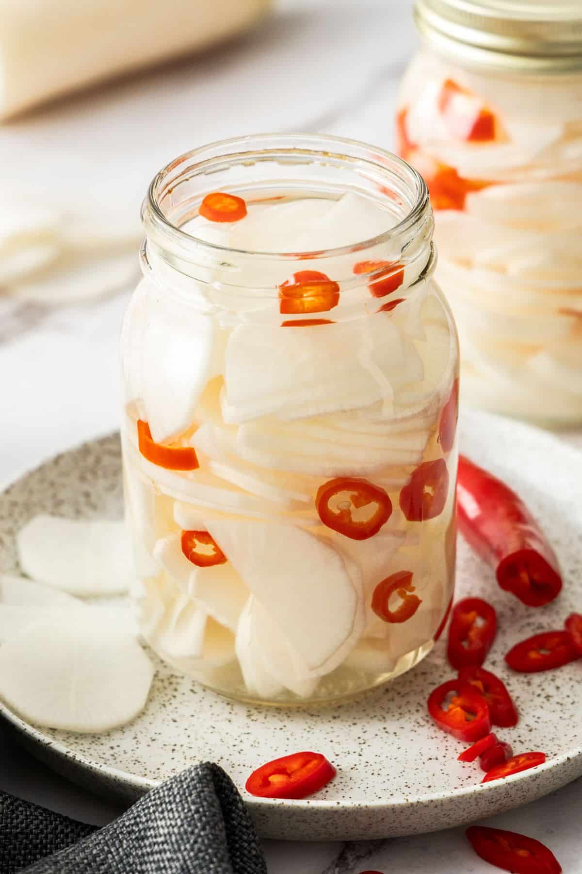 Glass jar of Pickled Daikon, sitting on a round plate, with some pieces of chilli on the plate.