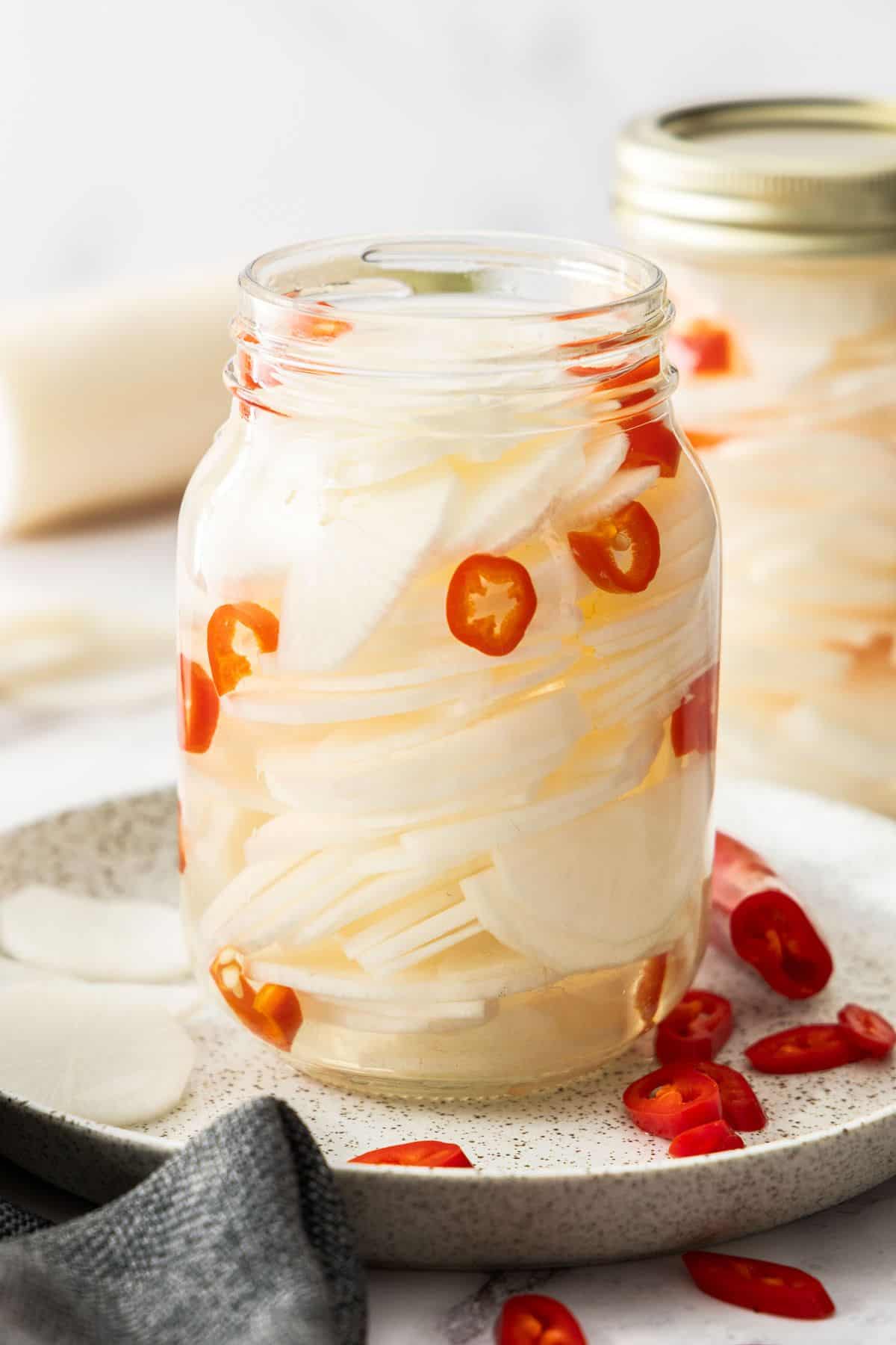 Glass jar of Pickled Daikon, sitting on a round plate, with some pieces of chilli on the plate.