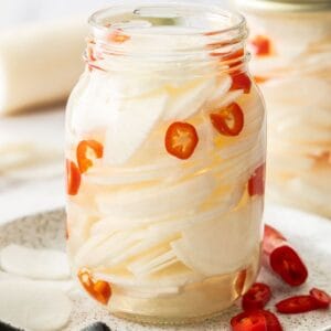 Glass jar of Pickled Daikon, sitting on a round plate, with some pieces of chilli on the plate.